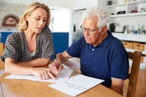 A mature woman assists an elderly man with estate planning in a paperwork at a kitchen table, in a bright, homey setting