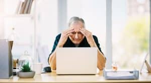 An elderly man appears stressed while using a laptop at a desk cluttered with papers and office supplies, presumably researching Medicaid's Look-Back Penalties.